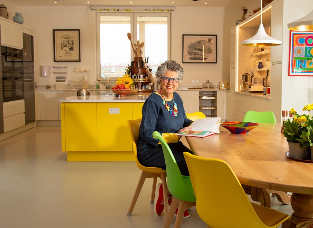 a woman in a blue dress sitting in a yellow handleless kitchen with a wooden dining table with yellow and green chairs