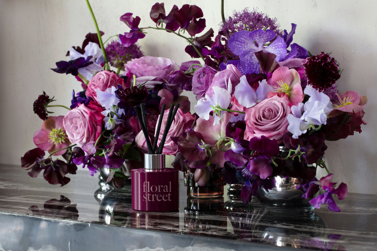 an arrangement of pink, maroon and purple flowers next to a burgundy perfume diffuser